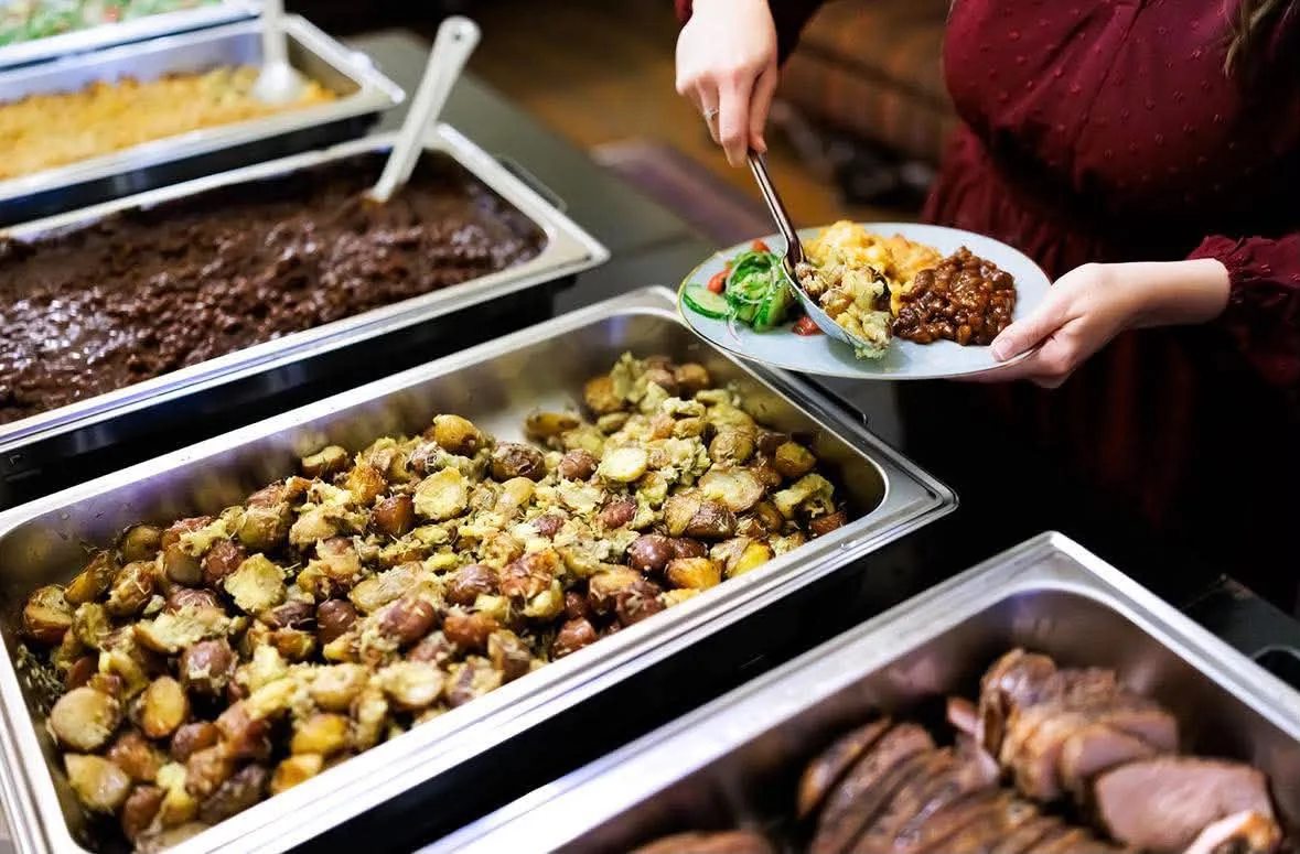A guest adding food to their plate beside trays of barbecue and sides.