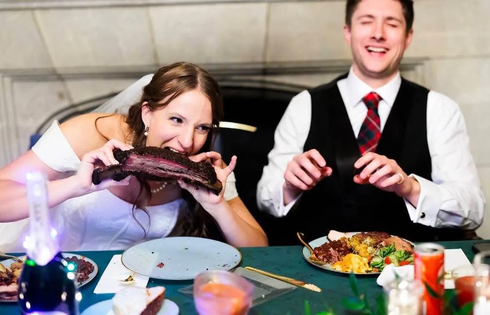 Bride biting into a barbecue rib while the groom smiles beside her.